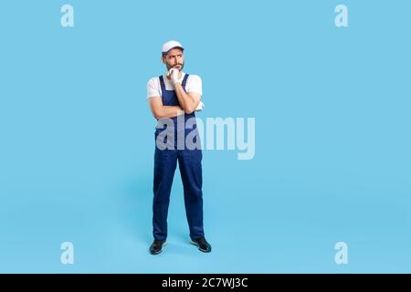 Full body pensive workman in overalls, cap and protective gloves standing, thinking intensely, pondering decision and having doubts. Profession of ser Stock Photo