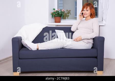 young beautiful pregnant woman using laptop and talking by phone at home Stock Photo