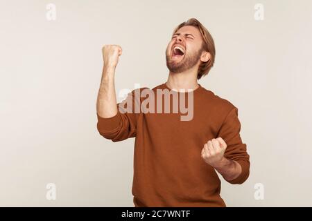 Joy of success, triumphal victory. Portrait of enthusiastic happy delighted man with beard in sweatshirt raising hands and screaming of euphoria. indo Stock Photo