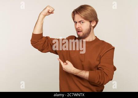 Look, how strong I am! Portrait of confident proud bearded man in sweatshirt demonstrating power in hand, pointing biceps, feeling energy to win succe Stock Photo