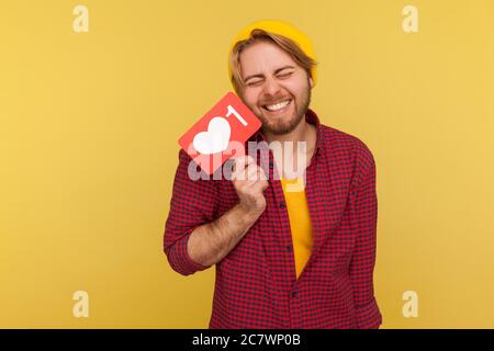 Extremely excited hipster guy in checkered shirt holding Heart Like counter button, smiling with pleasure expression, rejoicing popularity on Internet Stock Photo