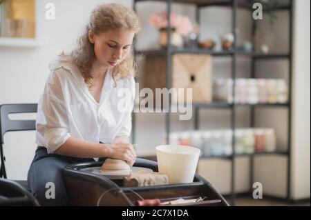 Young woman potter in apron at work. Craftsman artist shapes the jug with her hands and a special tool on pottery wheel. Stock Photo