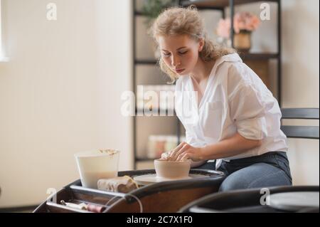 Young woman potter in apron at work. Craftsman artist shapes the jug with her hands and a special tool on pottery wheel. Stock Photo