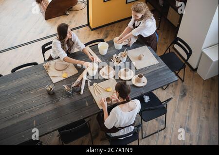 Top view of female potters during the working process in the clay workshop. Woman masters prepare ceramic and clay products at big wooden table Stock Photo