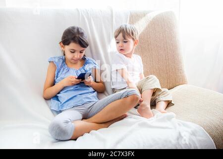 Children at home sitting on sofa, playing with tablet. Cute siblings brother and sister have a good time at home. Kids play a game on a cell phone Stock Photo