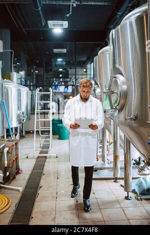 Full lenght of caucasian male professional technologist expert in white suit operating production machine in food factory and doing quality control an Stock Photo