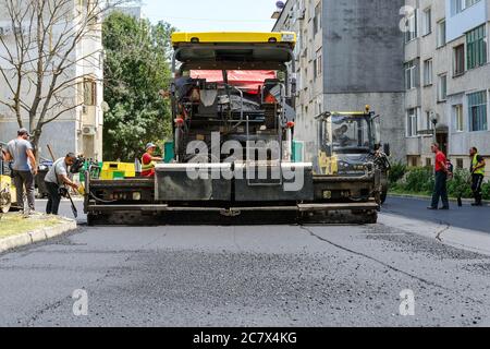 Varna, Bulgaria, July 16, 2020. Workers operating asphalt paver machine during road repairs in front of a multi-storey residential building on a day. Stock Photo