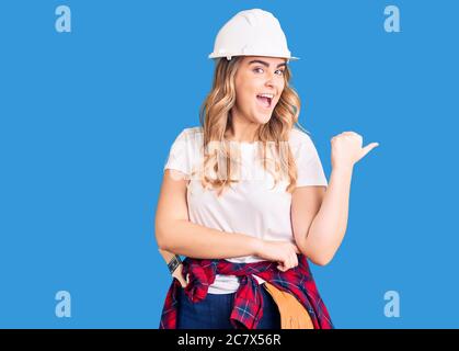 Young caucasian woman wearing security helmet smiling with happy face looking and pointing to the side with thumb up. Stock Photo