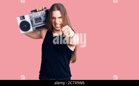 Young adult man with long hair wearing black clothes and holding boombox annoyed and frustrated shouting with anger, yelling crazy with anger and hand Stock Photo