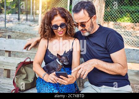 Middle age couple smiling happy using smartphone. Sitting on the bench with chihuahua dog at park Stock Photo