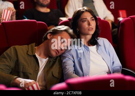 Portrait of young couple in cinema, focus on woman watching movie with bored boyfriend sleeping on her shoulder, copy space Stock Photo