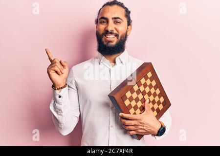 Young arab man holding chess smiling happy pointing with hand and finger to the side Stock Photo