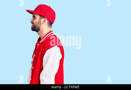 Young handsome man with beard wearing baseball jacket and cap looking to side, relax profile pose with natural face with confident smile. Stock Photo