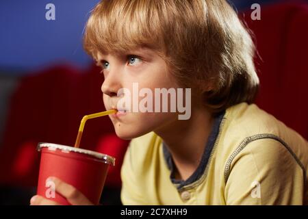 Head and shoulders portrait of cute blonde boy drinking soda while watching cartoons in cinema looking up at screen Stock Photo