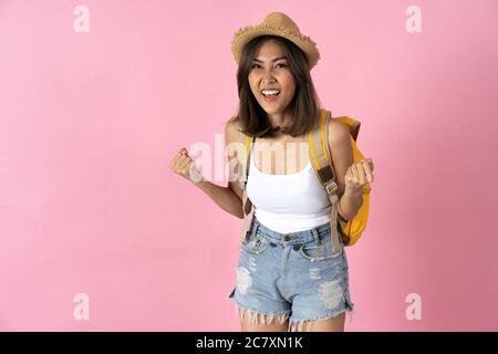 Young woman going on vacation wearing hat and backpack and clenching fists in excitement against plain pink background Stock Photo