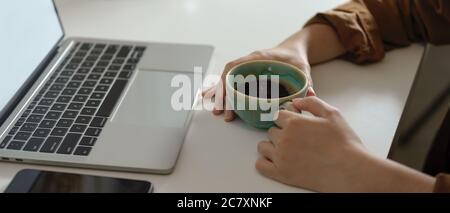 Side view of female holding coffee cup while looking on mock up laptop on white table with smartphone Stock Photo