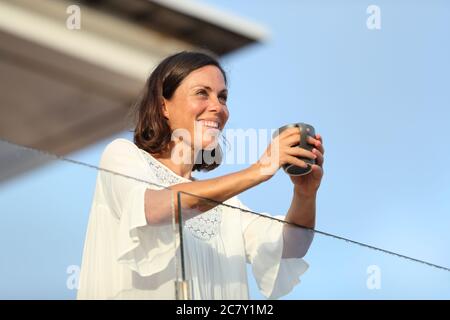 Happy adult woman with coffee cup looking away on a hotel balcony on vacation at summer Stock Photo