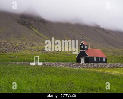 Saurbaejarkirkja black wooden red roof church on green grass field, steep hills in fog, Raudisandur, iceland west fjords