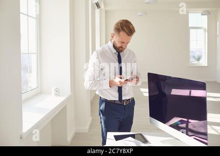 A bearded businessman uses a mobile reads sms text while standing in the workplace at the office. Stock Photo