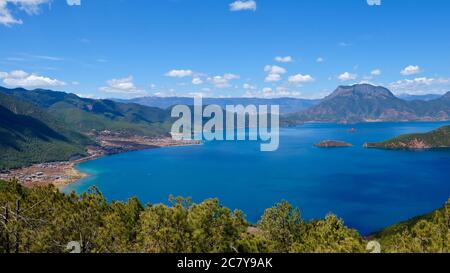 blue lake surrounded by green mountains. Green trees foreground. Sunny blue sky with white clouds. high angle view of Lugu lake in Yunnan China Stock Photo