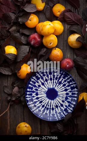 yellow ripe plums and empty ceramic plate among leaves on rustic old wooden table background, nobody Stock Photo