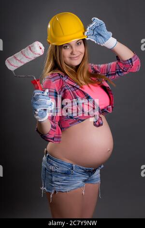 young beautiful pregnant woman in builder's helmet with  paint brush over grey background Stock Photo