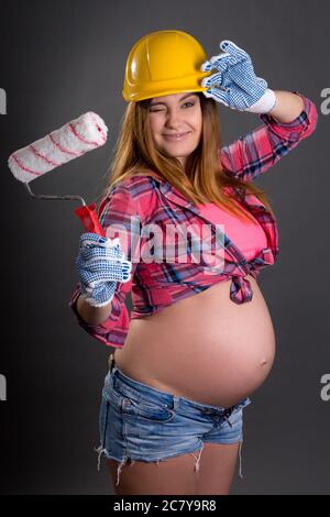 funny young beautiful pregnant woman in builder's helmet with  paint brush over grey background Stock Photo