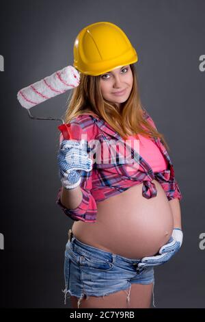 happy young beautiful pregnant woman in builder's helmet with  paint brush over grey background Stock Photo