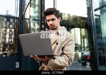 Young stylish man in trench coat confidently working on laptop on street Stock Photo