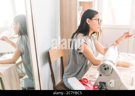 Young dressmaker woman sews clothes on sewing machine. Smiling seamstress and her hand close up in workshop. Focus on sewing machine and tissue. Stock Photo