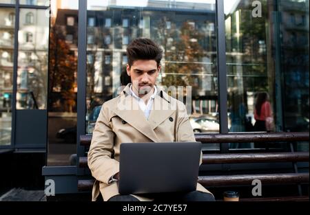 Young attractive stylish man in trench coat sitting on bench thoughtfully working on laptop on street Stock Photo