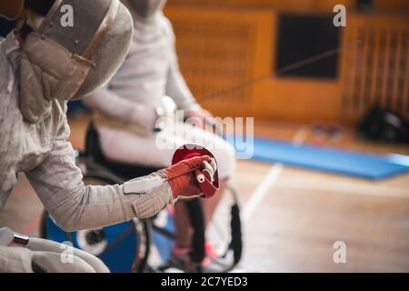 Close-up of man fencer hends wearing white fencing costume and holding his fencing mask and a sword on sunny background Stock Photo