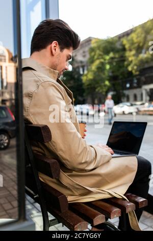 Back view of young stylish businessman in trench coat sitting on bench confidently working on laptop on street Stock Photo