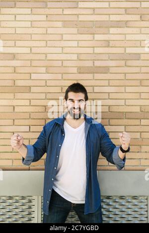 Joyful young bearded male in casual blue shirt posing on bricked wall background. Stock Photo