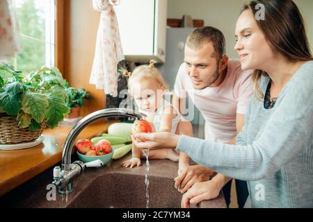 Young beautiful parents and her cute curly toddler daughter washing vegetables together in a kitchen sink getting ready to cook salad for lunch Stock Photo