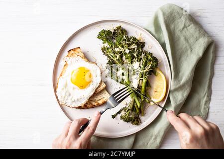 Roasted broccolini, lemon, fried egg with tost on the plate on the table decorated with napkin. Top view on healthy food. Female's hands with cutlery. Stock Photo