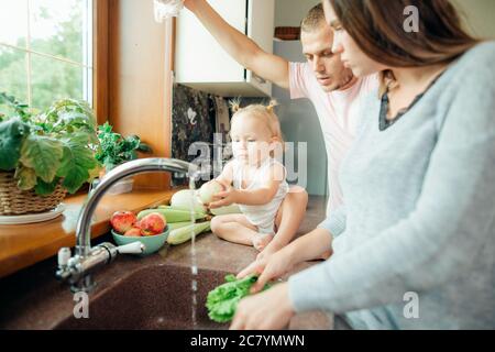 Young beautiful parents and her cute curly toddler daughter washing vegetables together in a kitchen sink getting ready to cook salad for lunch Stock Photo