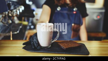 Barista wearing medical latex black gloves, making pink matcha latte with milk. Bartender preparing tasty drink and brownie cake Stock Photo