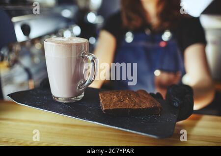 Barista wearing medical latex black gloves, making pink matcha latte with milk. Bartender preparing tasty drink and brownie cake Stock Photo