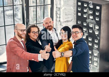 Happy business team. Group of cheerful business people in casual wear standing close to each other and showing their thumbs up Stock Photo