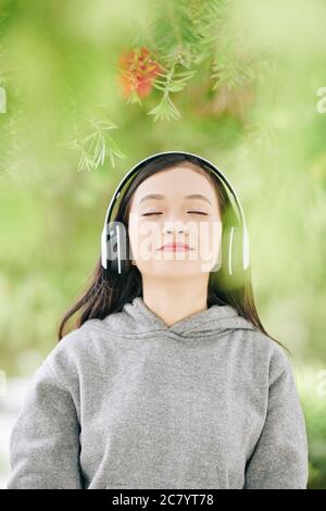Portrait of pretty teenage Asian girl in headphones closing eyes from enjoyment when listening to her favorite song in headphones Stock Photo