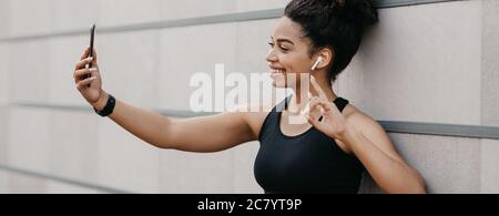 Muscular smiling african american girl in sport bra makes sign the peace and make photo on smartphone Stock Photo