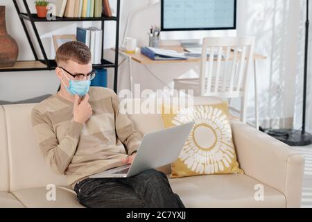 Pensive young software engineer in medical mask sitting on sofa at home and working on laptop Stock Photo