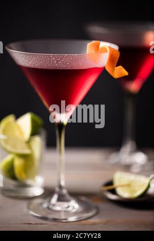 2 glasses of cosmopolitan cocktail garnished with a twist of orange and served with lime wedges on a dark rustic bar Stock Photo