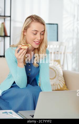 Pretty smiling young business lady eating slice of pizza when sitting on sofa at hme and working on financial report Stock Photo