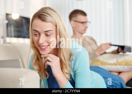 Smiling pretty young woman resting on sofa with her husband and watching tv series on tablet computer Stock Photo