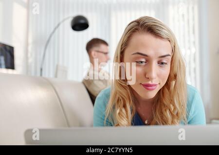 Pretty positive young woman lying on sofa and reading article on laptop screen Stock Photo