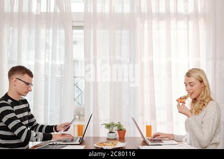 Young couple sitting at big table at home, eating pizza and working on laptops Stock Photo