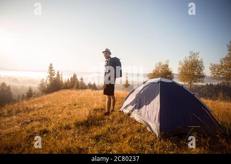 Handsome bearded man tourist exploring new places on top of mountains. Hiker man in hat with backpack happy to see autumn sunrise Stock Photo