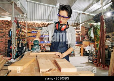 Young female carpenter in protective glasses polishing wooden plank using cordless sander Stock Photo